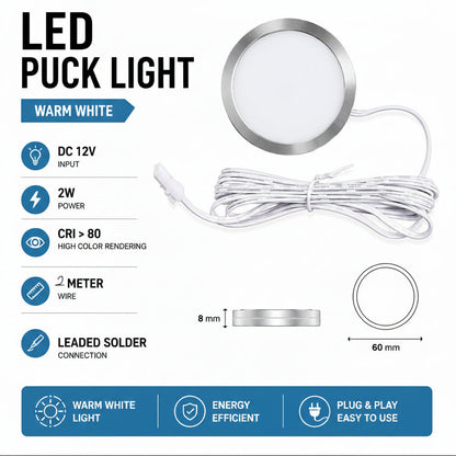 An LED puck light in warm white shown on a white background with product specifications, dimensions, and feature icons. The circular light has a silver metal rim and is shown with its attached white wire.